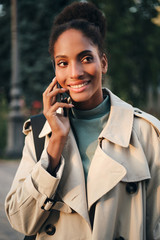 Beautiful casual African American girl in trench coat happily talking on cellphone outdoor