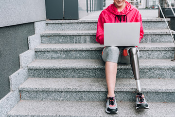 cropped view of smiling disabled sportswoman using laptop while sitting on stairs on street
