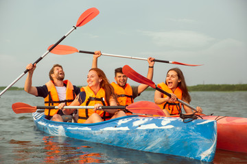 Happy young caucasian group of friends kayaking on river with sunset in the backgrounds. Having fun in leisure activity. Happy male and female model laughting on the kayak. Sport, relations concept.