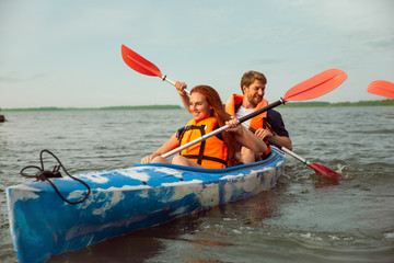 Happy young caucasian couple kayaking on river with sunset in the backgrounds. Having fun in leisure activity. Happy male and female model laughting on the kayak. Sport, relations concept. Colorful.