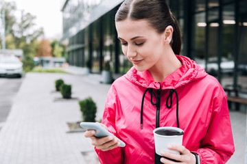 smiling sportswoman holding cup and using smartphone on street