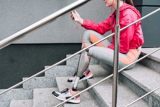 cropped view of disabled sportswoman holding smartphone and listening music on stairs