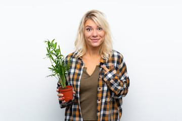 Young blonde gardener woman holding a plant over isolated white background with surprise facial...