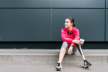 disabled sportswoman listening music in earphones and holding cup on street