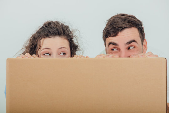 Funny wife and husband hiding behind the big carton box, together, looking at one another.