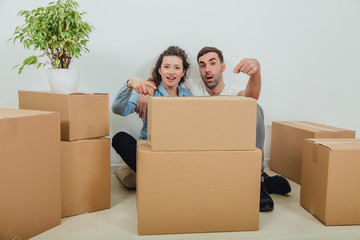 Funny wife and husband playing around, sitting behind the big carton boxes, pointing fingers at them, looking foolishly.