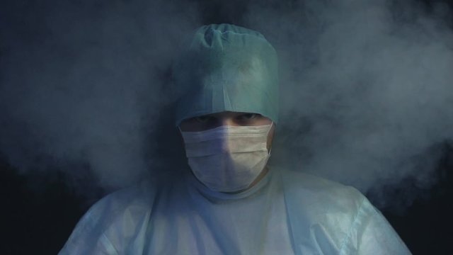 Surgeon looking at the camera in smoke in slow motion. Doctor is ready to start cut a patient's tissues in hospital. Save a life or kill concept. Surgery and operating table.