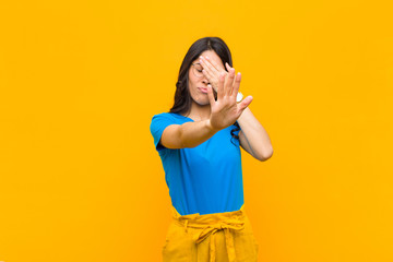 young pretty latin woman covering face with hand and putting other hand up front to stop camera,...