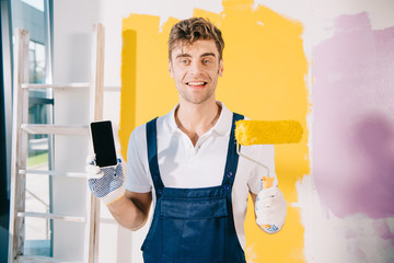 cheerful painter in uniform holding smartphone with blank screen and paint roller while smiling at...