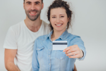 Blurred husband is hugging his wife. Pretty wife is holding a credit card in front of the camera. They are smiling. Focus on the card.