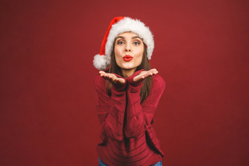 Christmas Santa hat woman portrait. Smiling amazed happy girl isolated against red background. Free copyspace for text. New year concept. Sending kiss.