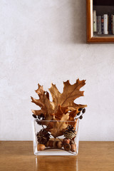 Still life with autumnal home decor of golden and brown dry leaves, cones and acorns in modern glass vase on wooden table, on wall background
