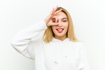 young blonde woman smiling happily with funny face, joking and looking through peephole, spying on...