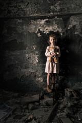 Obraz na płótnie Canvas sad kid standing and holding teddy bear in dirty room, post apocalyptic concept