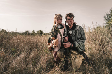 handsome man holding gun near kid with soft toy in field, post apocalyptic concept