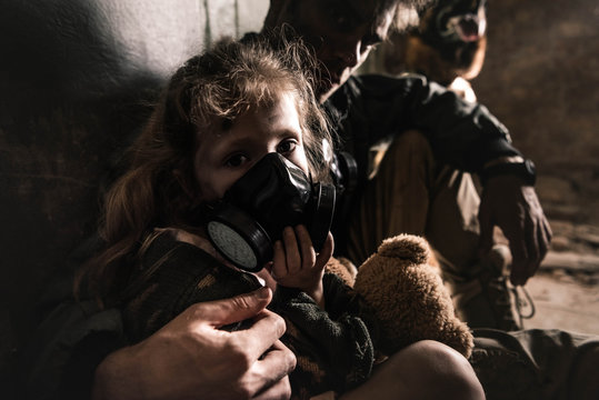 selective focus of man hugging kid in gas mask with teddy bear, post apocalyptic concept