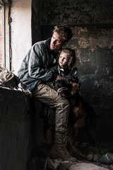 man hugging dirty child near german shepherd dog in abandoned building, post apocalyptic concept
