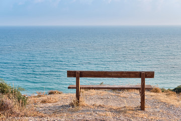 Fototapeta na wymiar Close-up shot of a wooden bench standing on a mountain at the beach of azure mediterranean sea and surrounded by a beautiful nature of Cyprus.