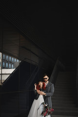 Fototapeta premium Beautiful couple of lovers hugging outdoors during wedding photoshoot. Slim young woman with red hair in modish leather jacket and wedding dress and handsome bearded man in the coat.