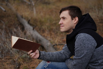 A young handsome man in casual clothes is sitting on a log and reading a book in the autumn forest.
