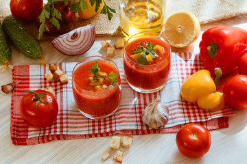 Tomato soup gazpacho in glass with herbs and ingredients on the kitchen. A beautiful summer soup spilled in glasses surrounded by juicy vegetables.