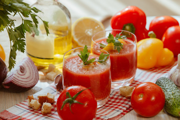 Tomato soup gazpacho in glass with herbs and ingredients on the kitchen. A beautiful summer soup spilled in glasses surrounded by juicy vegetables.