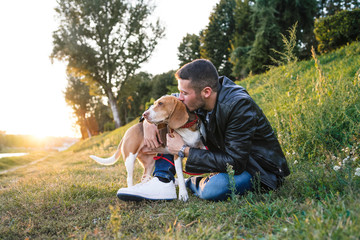 Young man sitting on the grass embraces the beloved dog at the park at sunset - Millennial in a...