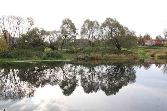 Autumn trees reflecting in calm water wiver with background of village houses and blue with clouds sky