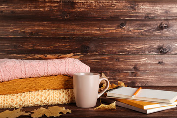 Folded sweaters with autumn leafs, cup of tea and notepads on brown wooden table