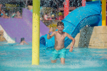 European boy splashing in pool at water park against colorful plastic slides, he is happy to spend...
