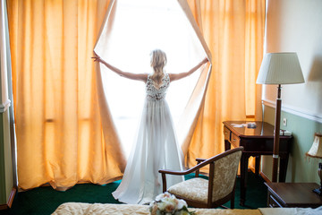 The blonde bride in a boudoir dress opens the curtains in the hotel room and looks out the window while preparing for the wedding and waiting for the groom. Inspiration, love, feelings, happiness.