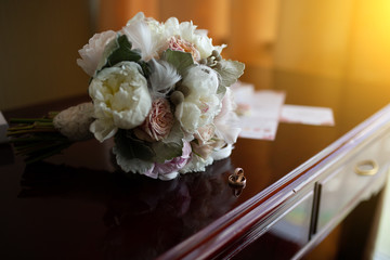 Wedding bouquet of peonies and roses lying on a wooden vintage table near wedding rings on the background of the invitation in the apartments to prepare the bride and expectation of the groom.