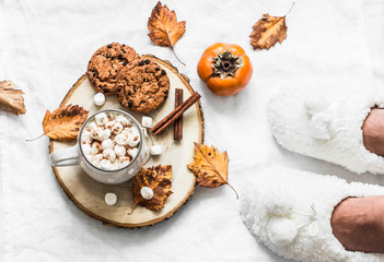 Cozy autumn still life - hot chocolate with marshmallow, cinnamon and chocolate chips cookies on the bed and women's feet in warm slippers on a light background, top view