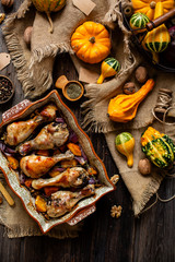 overhead shot of homemade tasty baked chicken drumsticks with pumpkin and red onion slices in baking ceramic pan stands on rustic wooden table with assorted mini pumpkins on sackcloth