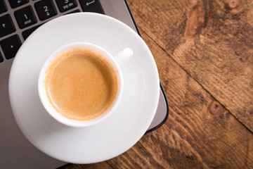 Cup of coffee on a laptop keyboard. Work place modern. Laptop (notebook) with cup of coffee and notepad with pen on old wooden table.