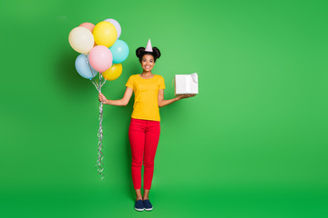 Full size photo of pretty dark skin lady holding air balloons big giftbox came to birthday party best guest wear casual yellow t-shirt red pants paper cap isolated green background