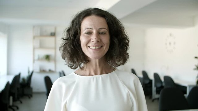 Cheerful middle aged Caucasian woman looking at camera. Front view of beautiful mature woman wearing white blouse standing at office. Female beauty concept