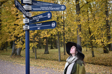 young beautiful pretty lady woman posing in a long green and brown dress gown overcoat walks down a lonely alley in the garden and looks at the information sign with emotions