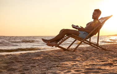 Young man reading book in deck chair on beach