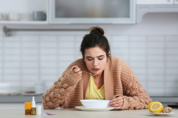Sick young woman eating soup to cure flu at table in kitchen