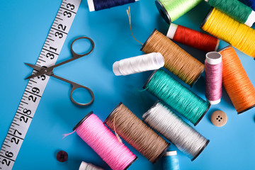 Sewing kit: scissors, measuring tape, thimbles, threads and buttons. Composition with threads and sewing accessories on blue background Colored thread coils on blue background, sewing, place for text