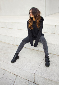 young casual brunette girl with long legs in sports black hoodie and jeans is sitting outdoor on the big white minimal stairs background and looking away, sport lifestyle concept, free space