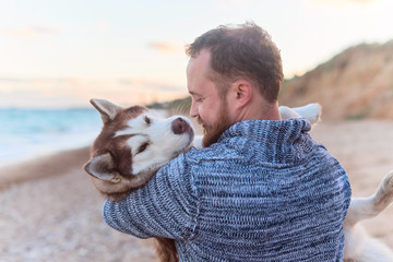 Man with beard in sweater holds a Siberian husky dog ​​on the background of the sea © iwavephoto