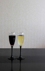Two wineglasses with white and red wine on black glass table. Gray blurred wall behind. Background with dark and light liquids in goblets and place for text.