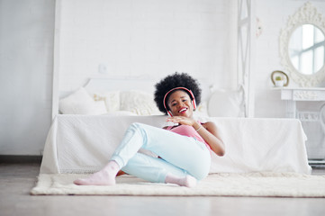 Young african american woman in pink singlet listen music on earphones at her white room.