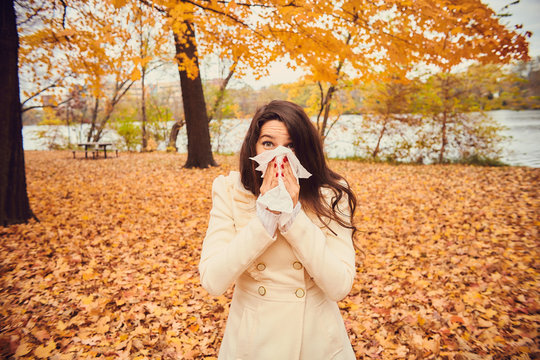 Portrait of young woman blowing nose in autumn park. Girl holding tissue, sneezing or coughing. Cold, flu, allergy