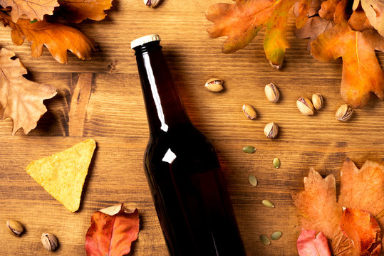 A bottle of dark beer, snacks and dry oak autumn leaves on a wooden background, top view. Autumn background with beer, nachos and pistachios flat lay. - Powered by Adobe