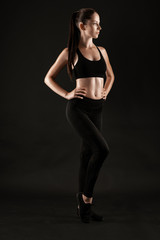 Fototapeta na wymiar Brunette woman in black leggings, top and sneakers is posing against a black background. Fitness, gym, healthy lifestyle concept. Full length.