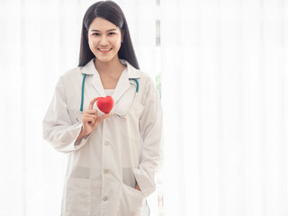 Portrait of a happy woman doctor standing and holding a red heart. Beautiful doctor smiling in a hospital or clinic.