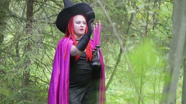 young witch with red hair and a black hat and a raspberry cloak conjures with candles in the forest.